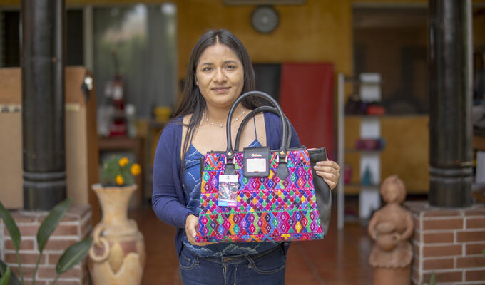 Guatemalan woman in centre holding large colourful/pink handbag, background has two brick bases and pillars of a shopfront
