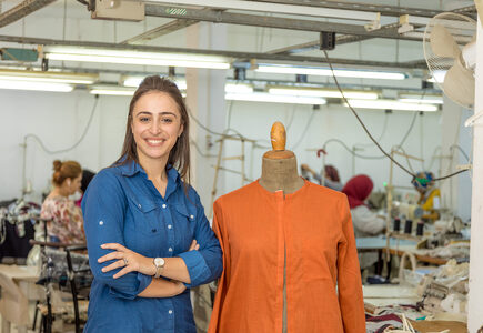 Une jeune femme souriante debout, les bras croisés à côté d'un mannequin de couturière portant une chemise orange vif. L’arrière-plan représente une usine, avec des femmes et des vêtements sur des étagères.