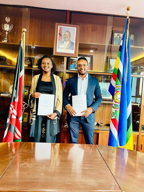 A man and woman in business attire hold signed agreements, standing between Ethiopian and Kenyan flags. 
