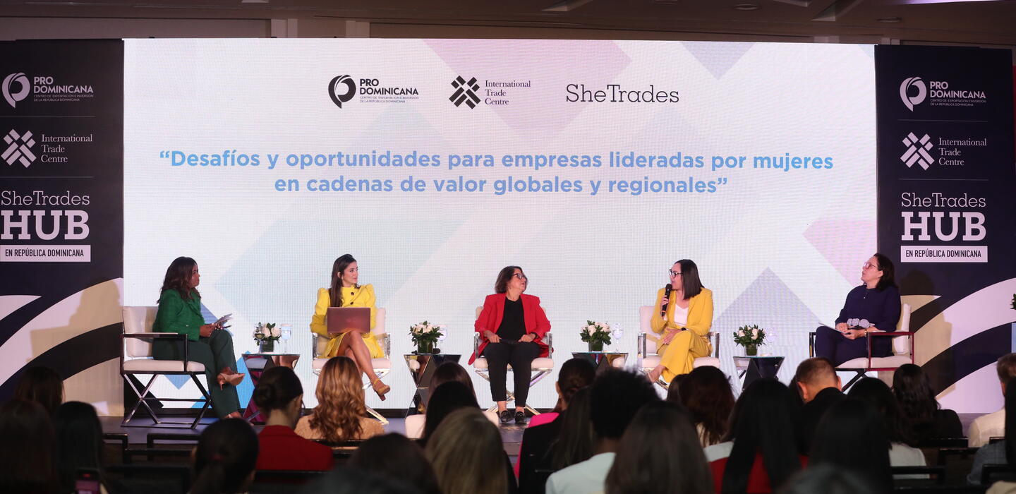 Group of panelists sit on stage under screen that reads SheTrades