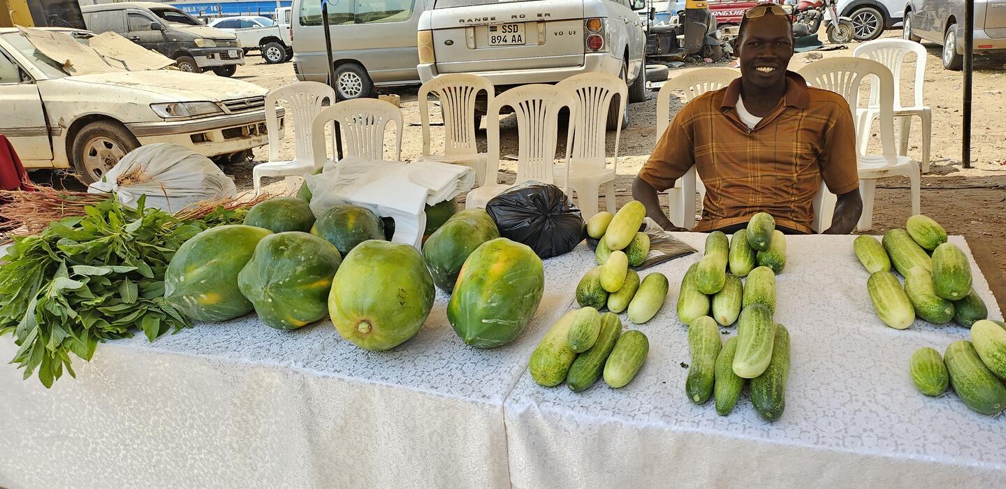 A South Sudanese farmer sits behind a table displaying his fruits and vegetables at a trade fair.