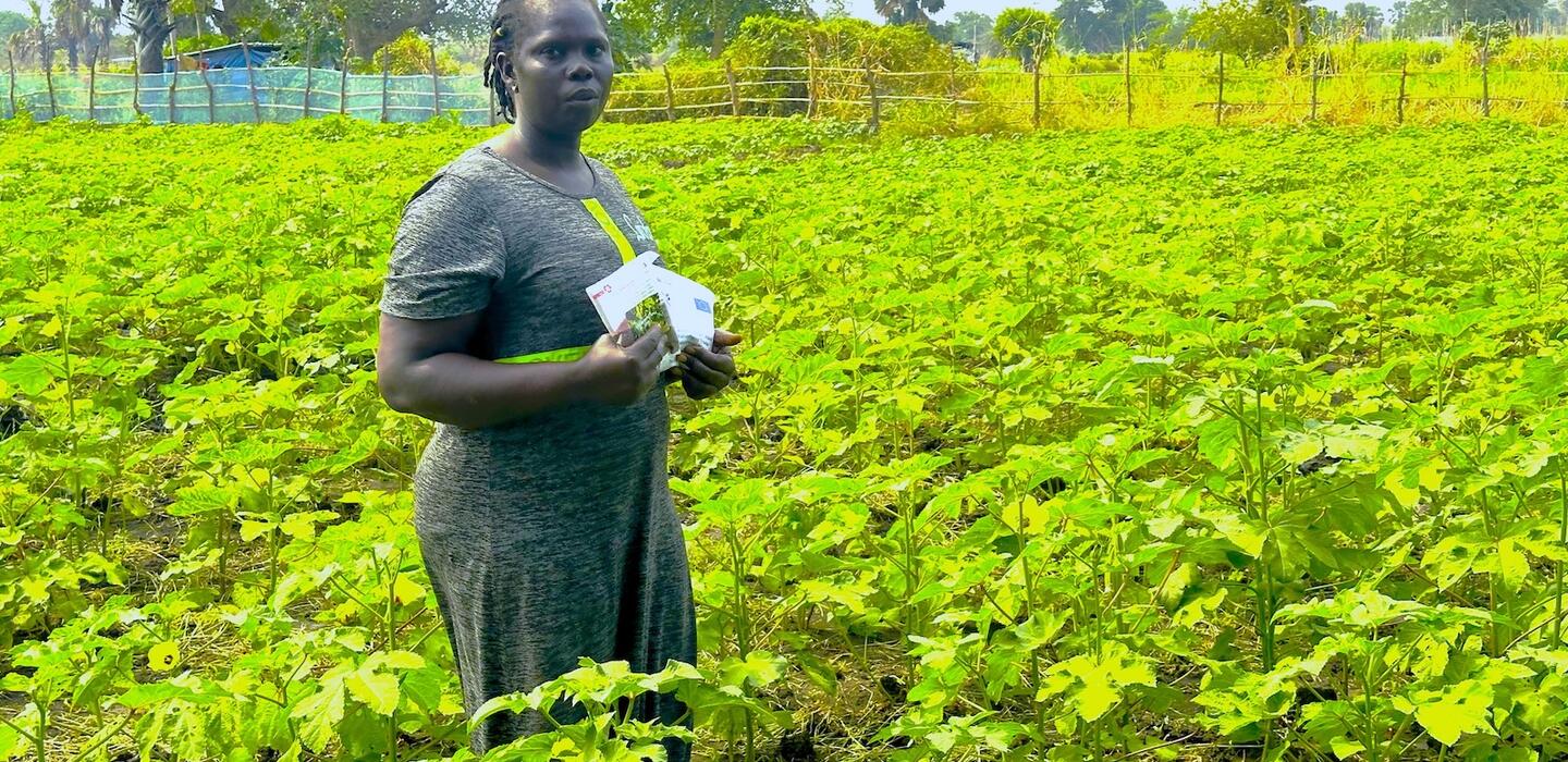 A woman South Sudanese farmer is standing in an okra field with 2 packets of seeds in her hands